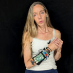 Perforamnce artist Avital Meshi stands in front of a black background. She has long blond hair draped over her right shoulder. She hold her right hand up over her chest, her left hand clasping the right. On her right forearm an electronic device is strapped. The photograph shows the electronics, uncovered.