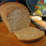 A loaf of pumpernickel rye bread sits on a wood cutting board. The end of the loaf lies in front of the cut loaf, which shows the rich and coarse texture of the bread. A bread knife sits next to the loaf.