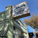 An upward tilted frame showing a sign with the words "California Typewriter" with a stylized image of a typewriter keyboard. The building upon which the sign hangs is a pale green, and the words "CALIFORNIA OFFICE MACHINES" is on the front above a showroom window.