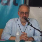 Jame Mustich sits behind a microphone in a blue Oxford shirt. A water bottle sits in front of him. He has his hands folded as he talks, probably at a conference or a meeting.