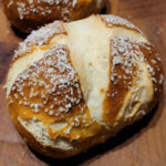 A closeup of a Laugenbrötchen, which is a dark brown roll, with a croscut on top and covered with coarsely ground salt.