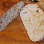 A loaf of bread with a freshly slived piece laying beside it. The bread crustis dark brown, and the slice shows light and airy center of the slice. It is a batard, basically a fat baguette.