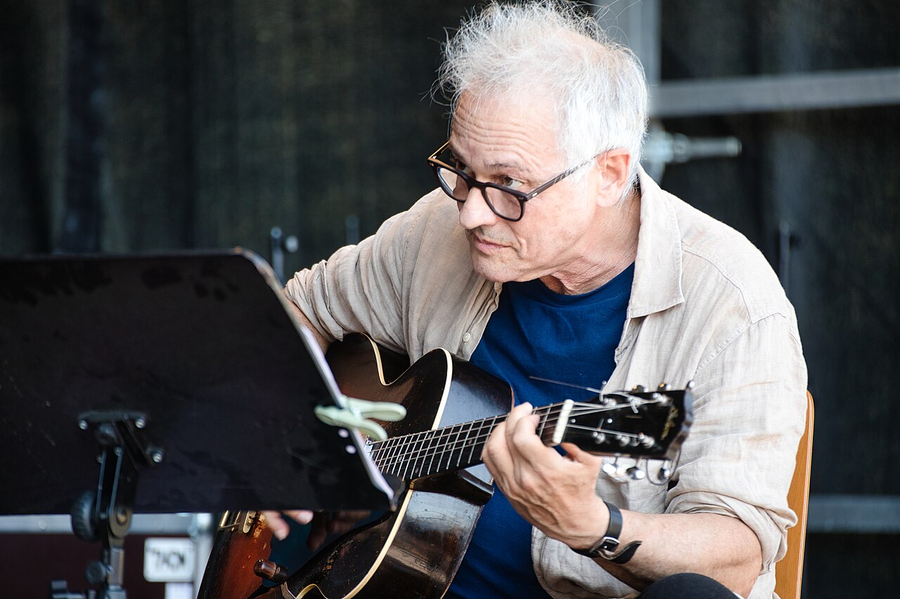 Marc Ribot leans over his guitar in his performance at INNtöne Jazzfestival 2024. His white hair os frizzy and he looks at a fellow musician over his glasses.