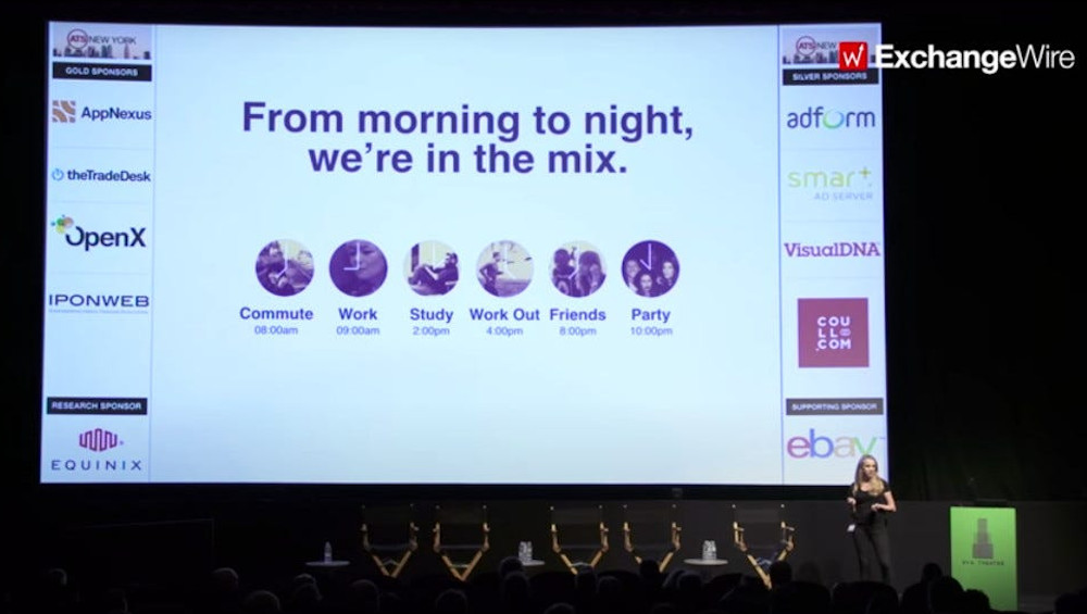 A darkened auditorium with a large screen showing a presentation slide. To the lower right, a woman in a long dress stands as she presents.