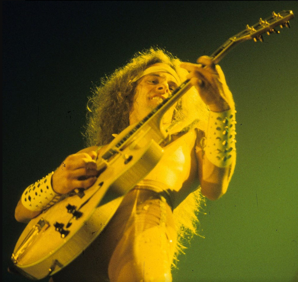 Ted Nugent when he was young, playing a guitar in yellow-colored stage lighting. His long light-colored hair flows from his head.
