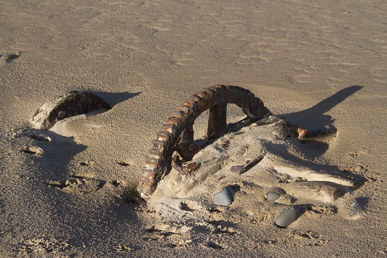 A very old and heavily rusted gear is nearly completely covered by fine sand, It is part of a long abandoned machine.